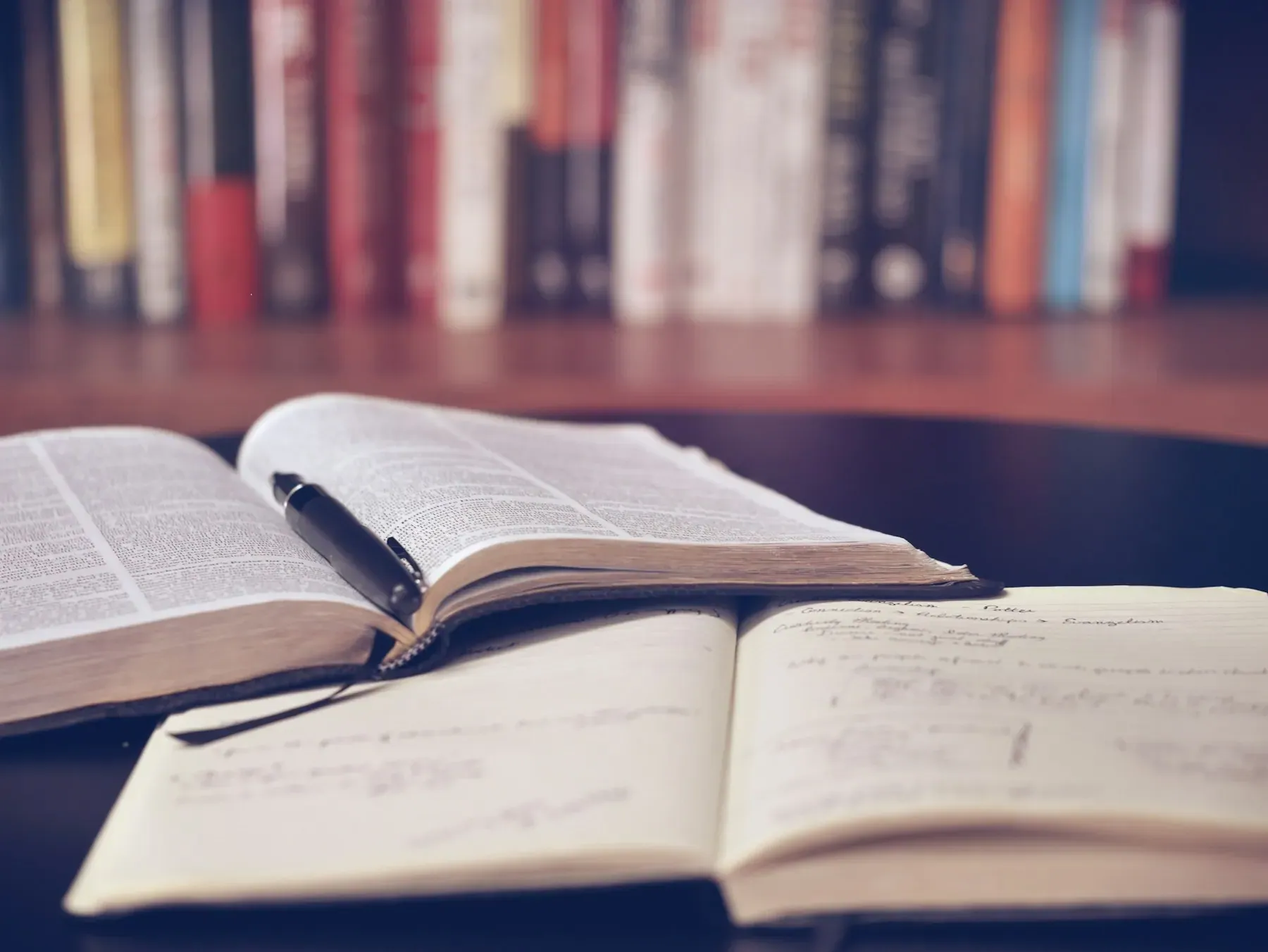Libro abierto sobre escritorio de madera, con estantería de libros al fondo desenfocada